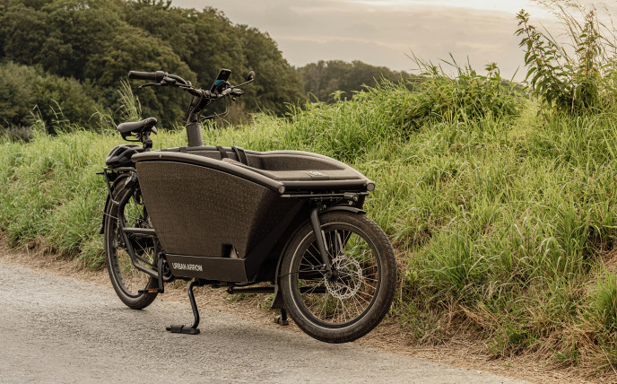 Ein schwarzes Lastenfahrrad ist auf einem unbefestigten Weg neben einer Wiese geparkt, mit Bäumen im Hintergrund unter einem bewölkten Himmel.