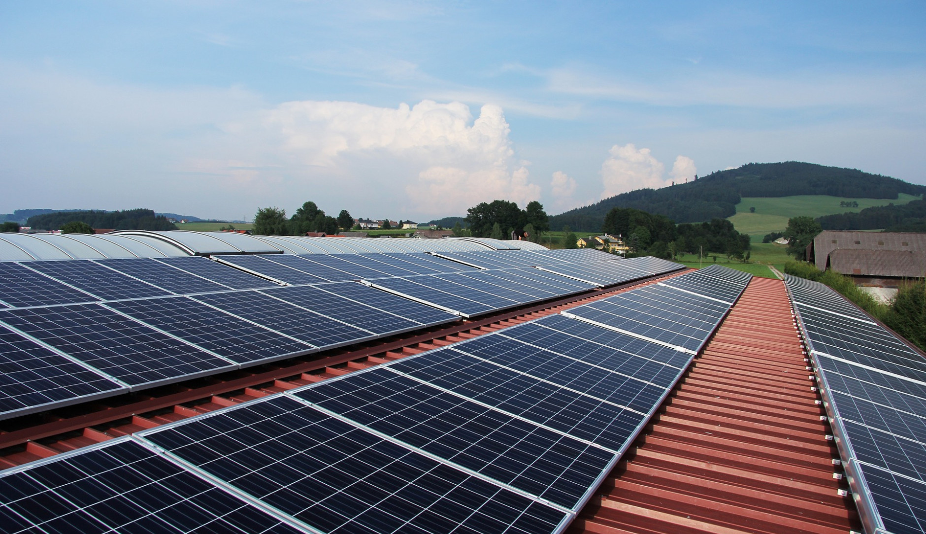 Das Bild zeigt Solarpaneele, die auf einem Dach installiert sind. Im Hintergrund sieht man eine hügelige Landschaft mit Bäumen und Häusern.