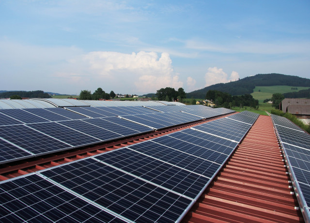 Das Bild zeigt Solarpaneele, die auf einem Dach installiert sind. Im Hintergrund sieht man eine hügelige Landschaft mit Bäumen und Häusern.