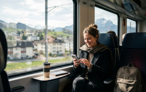 Eine junge Frau sitzt lächelnd am Fenster eines modernen Schweizer Zugs und bedient ihr Smartphone. Auf dem Tischchen vor ihr stehen ein Mehrwegbecher und ein Notizbuch, während draußen sanft verschwommene Gebäude und alpine Hügel im Tageslicht vorbeiziehen.