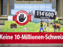Eine Pyramide aus bedruckten Boxen vor einer Steinfassade zeigt eine ländliche Schweizer Landschaft mit Bergen, einer Familie, einer Kuh und Schafen. Darauf integriert sind ein Stoppsymbol mit einer Hand und dem Text «10 Millionen» sowie die Schriftzüge «Nachhaltigkeits-Initiative:», «114'600 Unterschriften» und gross auf rotem Grund am Fuss der Pyramide «Keine 10-Millionen-Schweiz!».