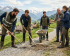 Fünf Personen in funktioneller Arbeitskleidung verrichten handwerkliche Arbeiten auf einem Schotterweg in einer alpinen Landschaft. Im Vordergrund links arbeiten zwei Männer mit Schaufel und Pickel am Boden. In der Mitte steht ein Mann hinter einer mit Erde und Steinen gefüllten Schubkarre. Auf der rechten Seite stehen eine Frau mit einer Harke und ein Mann im Gespräch vertieft. Im Hintergrund ist ein von grünen Wiesen umgebenes Dorf vor einer Kulisse aus Bergen mit schneebedeckten Gipfeln zu sehen.