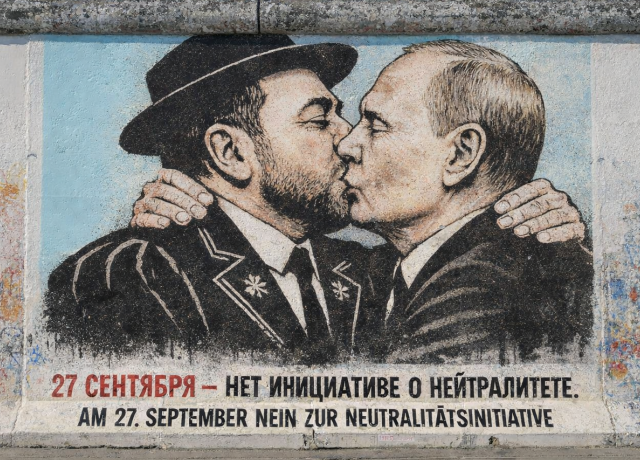 Ein Foto zeigt ein großes, farbenfrohes Wandgemälde auf einem verwitterten Betonmauerabschnitt, das eine Parodie des berühmten 