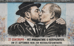 Ein Foto zeigt ein großes, farbenfrohes Wandgemälde auf einem verwitterten Betonmauerabschnitt, das eine Parodie des berühmten 