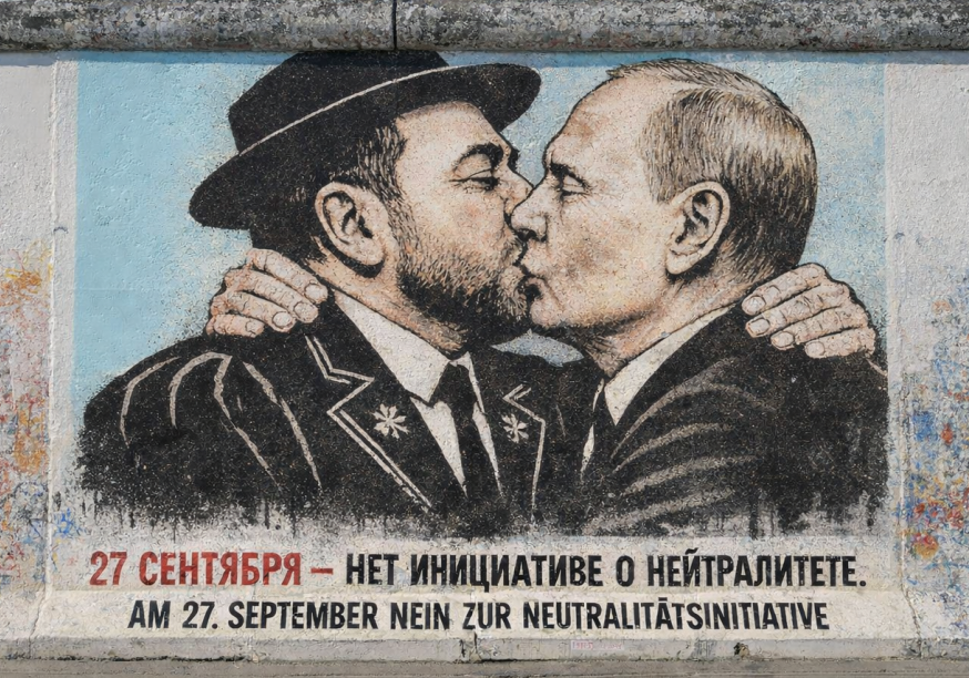 Ein Foto zeigt ein großes, farbenfrohes Wandgemälde auf einem verwitterten Betonmauerabschnitt, das eine Parodie des berühmten 