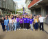 Eine Gruppe von etwa 20 gut gelaunten Menschen posiert im Freien auf einem asphaltierten Platz vor modernen Gebäuden. Mehrere Personen in der Mitte tragen violette T-Shirts oder Hoodies mit dem weissen Aufdruck «Volt». Einige Personen halten Blumen hoch oder haben die Arme jubelnd in die Luft gestreckt. Im Hintergrund ist eine violette Volt-Fahne zu sehen. Das Bild vermittelt eine positive, gemeinschaftliche Atmosphäre.