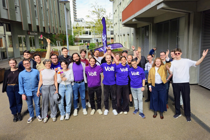 Eine Gruppe von etwa 20 gut gelaunten Menschen posiert im Freien auf einem asphaltierten Platz vor modernen Gebäuden. Mehrere Personen in der Mitte tragen violette T-Shirts oder Hoodies mit dem weissen Aufdruck «Volt». Einige Personen halten Blumen hoch oder haben die Arme jubelnd in die Luft gestreckt. Im Hintergrund ist eine violette Volt-Fahne zu sehen. Das Bild vermittelt eine positive, gemeinschaftliche Atmosphäre.